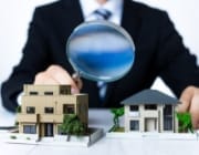 ビジネススーツを着た人物が虫眼鏡で住宅模型を詳しく調べている様子